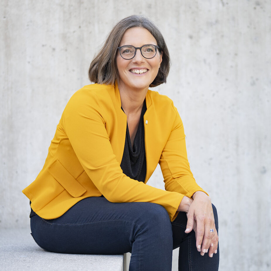 Frau mit rundlicher Brille und orangem Blazer sitzt auf einer Betonstiege