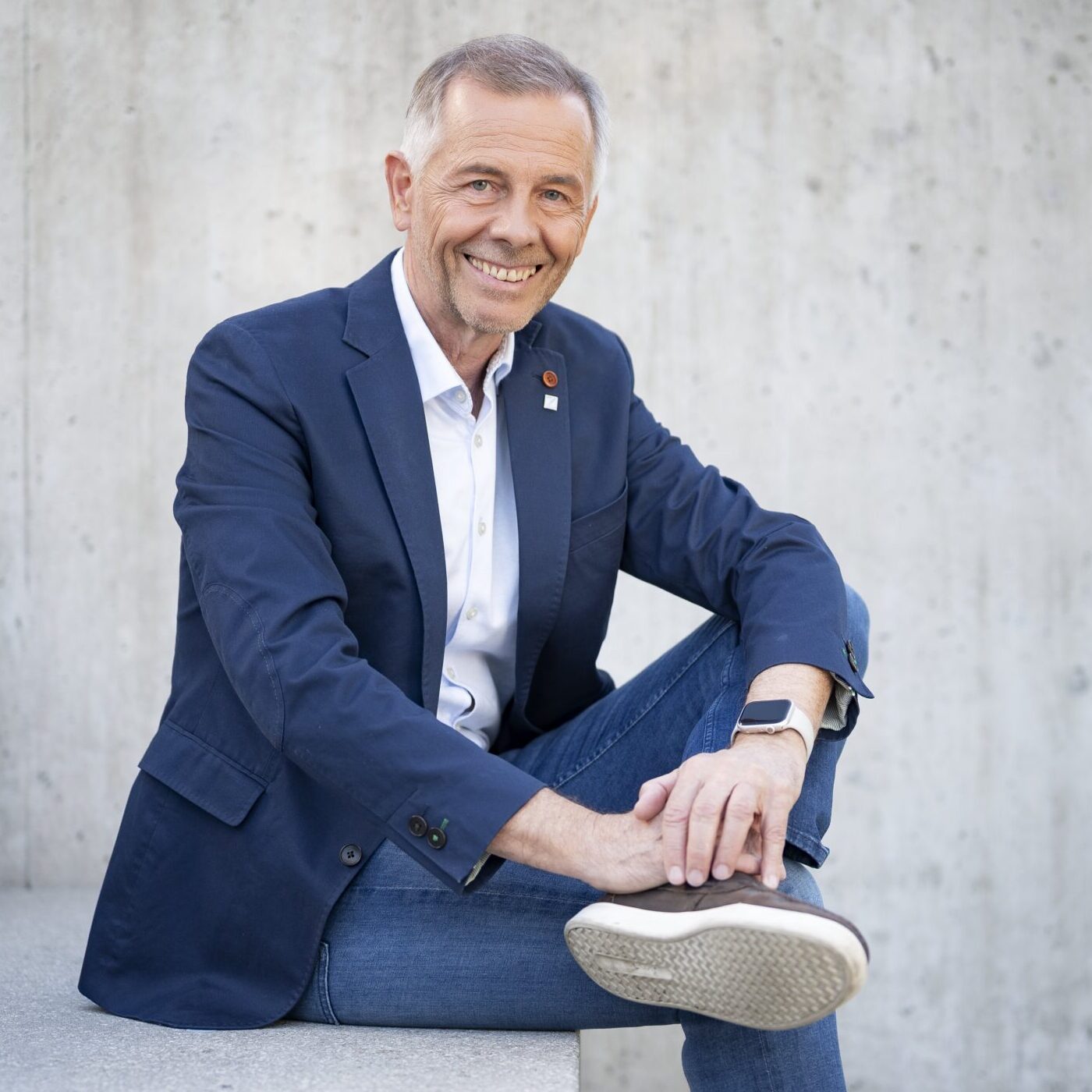 Mann mit grau-melliertem Haar und legerem dunkelblauem Sakko und Jean sitzt auf einer Beton Treppe