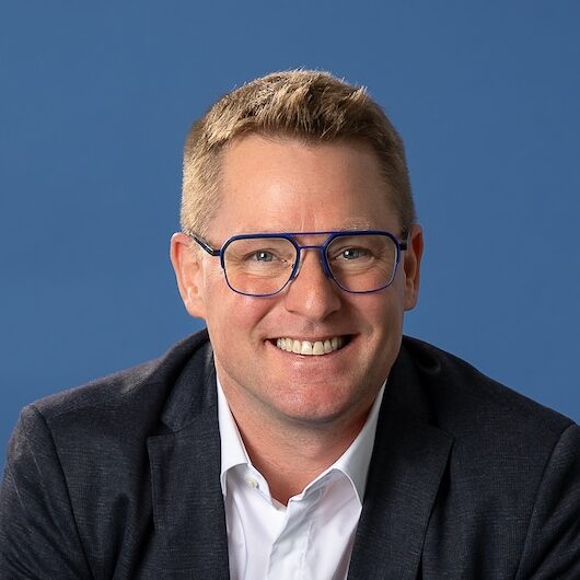 Portraitfoto eines Mannes mit blauer Brille, dunklem Sakko