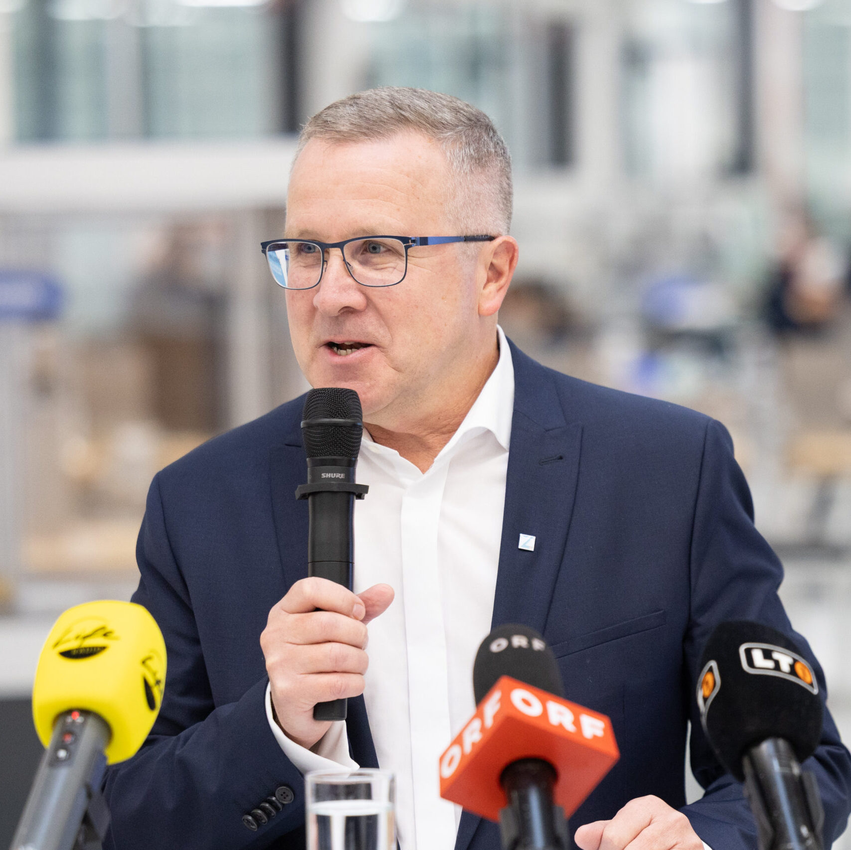 Ein Mann mit Brille und dunklem Anzug spricht in ein Mikrofon. Im Vordergrund sind weitere Mikrofone zu sehen.