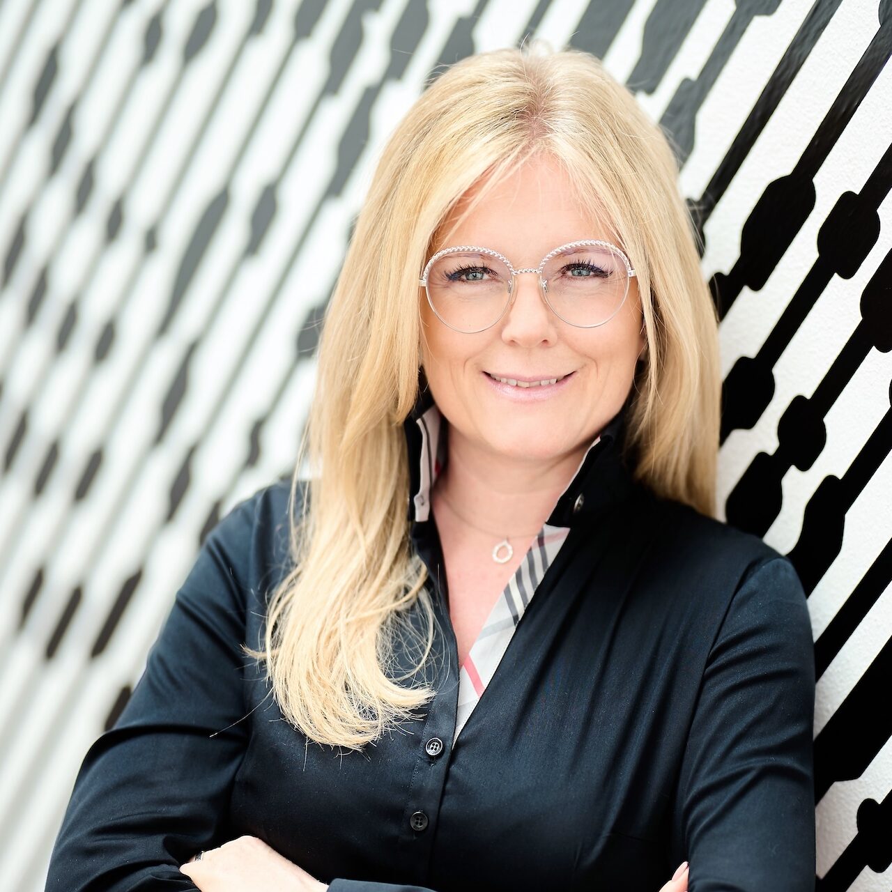 Portraitfoto einer Frau mit Brille und dunkler Bluse, angelehnt an eine Wand mit abstraktem Muster