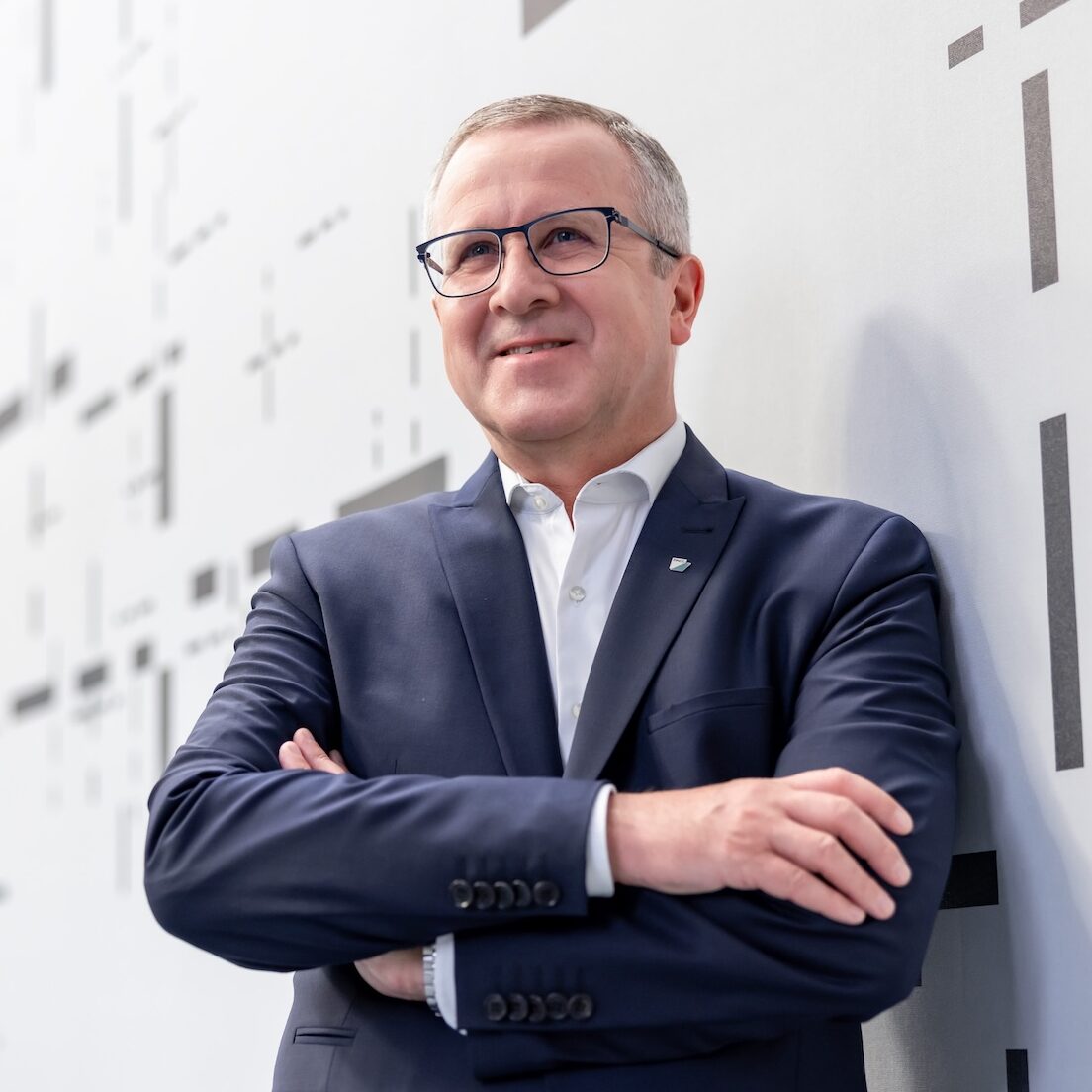 Portraitfoto eines Mannes im blauen Anzug mit weißem Hemd lehnt an einer Wand